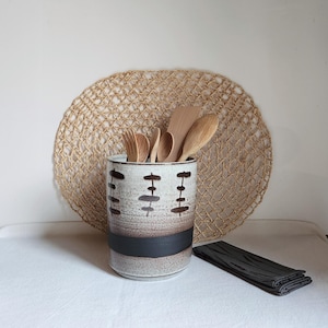 Könnte beinhalten: Ein Keramik-Utensilienhalter mit weiß-braun gesprenkelter Oberfläche und schwarzen geometrischen Designs. Er enthält Holzlöffel und eine Gabel. Ein gewebter Platzdeckchen und ein gefaltetes graues Tuch befinden sich im Hintergrund.