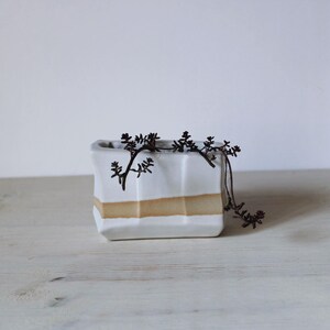 May include: A rectangular white ceramic planter with a horizontal tan band around the centre. The planter features a faceted, geometric surface texture and contains a small, dark-coloured trailing plant that hangs over the top edge.
