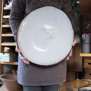 Op de afbeelding: Een grote, ronde, handgemaakte keramische kom met een glanzend wit binnenglazuur met zichtbare horizontale geribbelde textuurlijnen. De kom heeft een donkere, contrasterende rand en een gladde, brede komvorm.