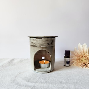 May include: A ceramic essential oil diffuser with a lit tea light inside. The diffuser is cylindrical with a textured, light grey and brown finish. A small bottle of essential oil and a flower are next to the diffuser.