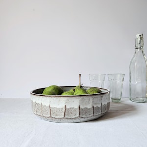 Op de afbeelding: Een ronde keramische kom met een gespikkelde grijze basis en een wit geglazuurde rand, gevuld met verschillende groene peren. Naast de kom staan twee heldere glazen bekers en een hoge, lege glazen fles met een metalen beugelsluiting.