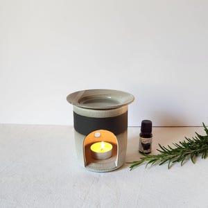 Op de afbeelding: Een keramische etherische olieverspreider met een zwarte band, een kleine kaars erin en een klein flesje etherische olie. De diffuser is grijs en wit, met een ronde bovenkant en een gebogen opening voor de kaars.