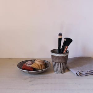 Könnte beinhalten: Eine braun-weiße Keramiktasse mit Make-up-Pinseln, eine kleine Schale mit Bürste und Seife und ein gefaltetes graues Tuch auf einer Holzoberfläche. Die Tasse hat vertikale Linien. Die Schale ist grau mit dunklem Rand.