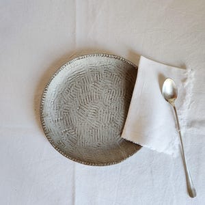 Peut inclure: Une assiette en céramique texturée, de couleur gris clair, avec un motif. Une serviette en lin blanche et une longue cuillère en argent sont placées à côté de l'assiette. L'ensemble est disposé sur une nappe blanche.