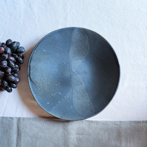 Op de afbeelding: Een ronde, donkerblauwe keramische kom met een matte afwerking en een subtiele, lichtere blauwe verticale glazuurstreep in het midden. Het oppervlak vertoont vage, gespikkelde textuurpatronen. Een tros donkerpaarse druiven ligt aan de linkerkant.