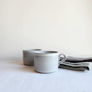 May include: Two ceramic mugs with a speckled light grey glaze and a smooth finish. One mug features a curved handle on the side, while the other is handleless. The mugs have a cylindrical shape with a slightly wider rim and a flat base.