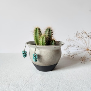 Può includere: Orecchini con perline turchesi appesi a un piccolo vaso di ceramica macchiato con un cactus all'interno. Il vaso ha una base nera e una parte superiore grigio chiaro. Gli orecchini sono fatti di piccole perline rotonde.