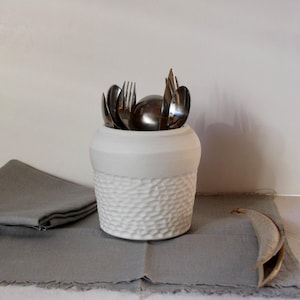 Op de afbeelding: Een witte keramische bestekhouder met een getextureerd, kuiltjesvormig onderste gedeelte en een glad bovenste gedeelte. De houder bevat diverse roestvrijstalen vorken, lepels en messen. De houder staat op een grijze linnen placemat.