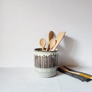 Puede incluir: Un porta utensilios cilíndrico de cerámica con un patrón de esmalte de goteo en blanco y marrón oscuro. Dentro del recipiente hay tres cucharas de madera clara y una espátula de madera. Un tenedor de madera descansa sobre una servilleta gris.