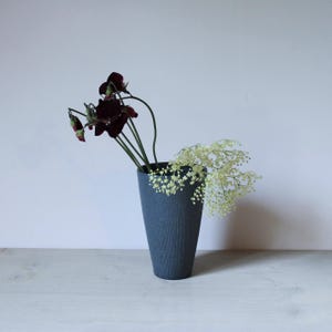 Puede incluir: Un jarrón de cerámica azul oscuro, de forma cónica, con una textura de finas estrías verticales. El jarrón contiene un pequeño arreglo de flores de color granate oscuro y una rama de flores de relleno delicadas, de color verde claro.