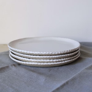 May include: Four white ceramic plates with a textured, scalloped edge are stacked. The plates rest on a grey surface. The plates have a simple, elegant design. The plates are suitable for serving food.