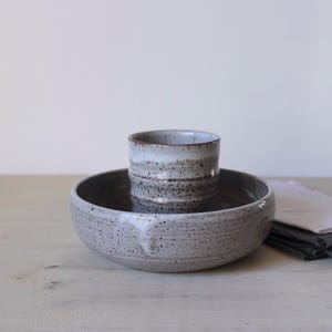 Puede incluir: Un juego de cuenco y taza de cerámica hechos a mano. El cuenco es ancho y poco profundo, con un acabado gris moteado. La taza es cilíndrica, con un esmalte blanco y marrón. El juego está diseñado para servir alimentos o bebidas.