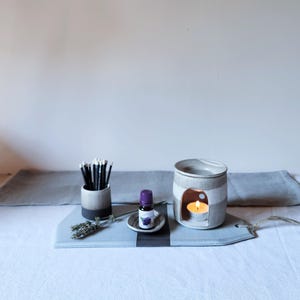 Op de afbeelding: Een keramische etherische olieverspreider met een brandende kaars, een klein flesje olie en een houder voor wierookstokjes. De diffuser is grijs en wit gestreept. De items zijn gerangschikt op een grijs en blauw dienblad.