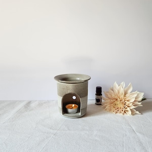 May include: A beige and grey ceramic essential oil diffuser with a lit tea light candle inside. The diffuser has a textured design. A small bottle of essential oil and a cream-coloured flower are next to the diffuser.