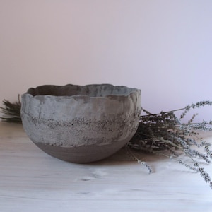 Peut inclure: Un bol en céramique grise fait à la main, avec un bord ondulé texturé et une bande de détails gravés. Le bol est posé sur une surface en bois blanc, avec des brins de lavande séchée. Esthétique neutre et terreuse.