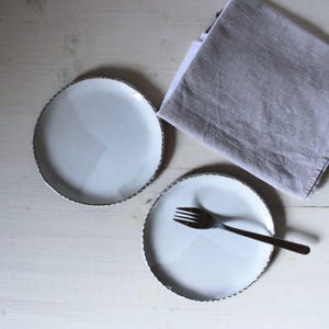 Peut inclure: Deux assiettes en céramique blanche avec un bord décoratif et une fourchette argentée sont disposées sur une surface en bois clair. Une serviette en lin grise pliée est également présente.