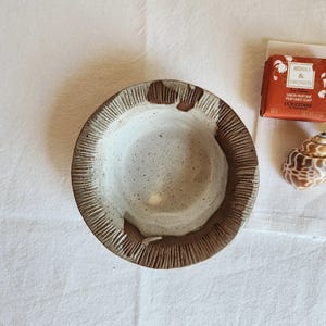 Peut inclure: Un bol en céramique fait à la main avec un bord texturé et un intérieur moucheté. Le bol est marron clair et blanc. Un pain de savon orange et un coquillage sont à côté du bol.