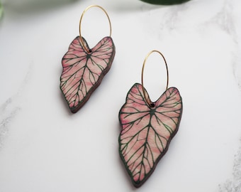 Puede incluir: Un par de pendientes de aro dorados con dijes en forma de hoja rosa y verde. Las hojas tienen un patrón de nervaduras detallado y un efecto de acuarela.
