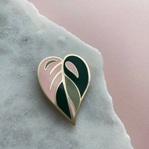 May include: A gold heart-shaped enamel pin with a pink and green leaf design.