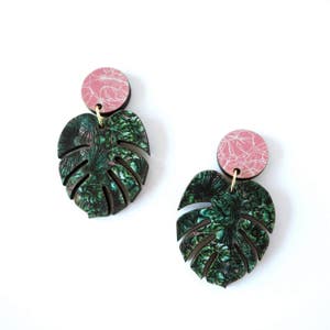 May include: A pair of earrings featuring a pink and green tropical leaf design. The earrings are made of wood and have a pink circle on top of the leaf.