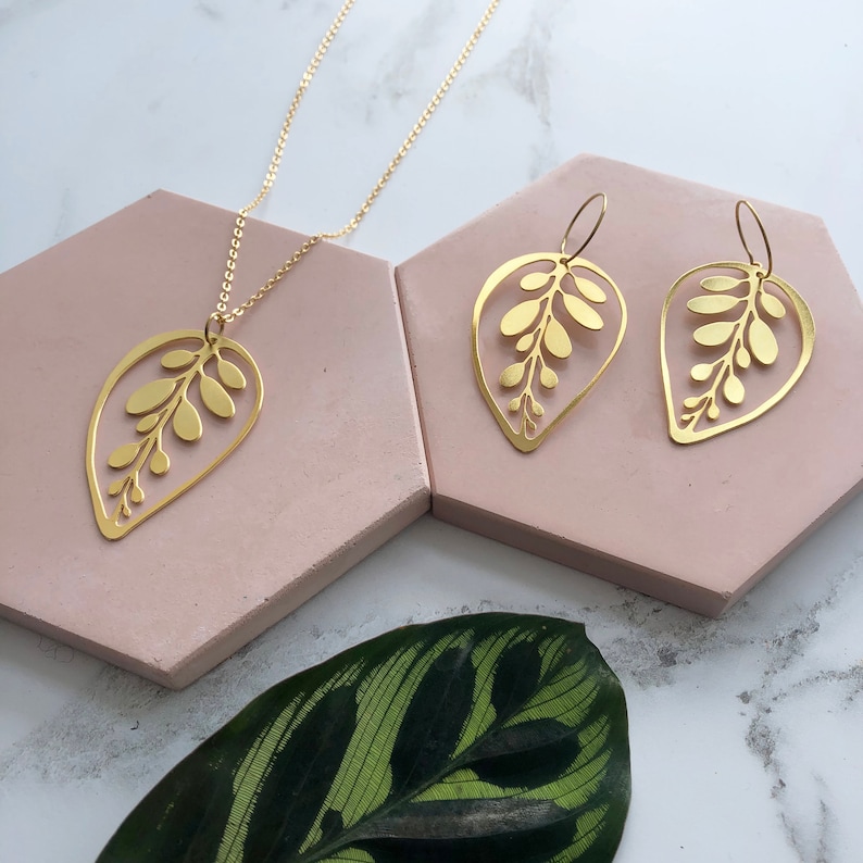 May include: A gold necklace and earrings set featuring a delicate leaf design with intricate cutouts. The jewelry is displayed on a pink surface with a green leaf in the foreground.