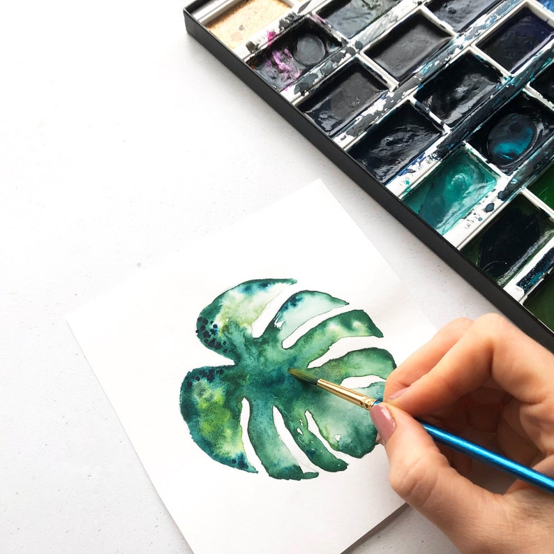 May include: A watercolor painting of a green monstera leaf being painted with a brush. The artist is using a blue and gold paintbrush to add details to the leaf.