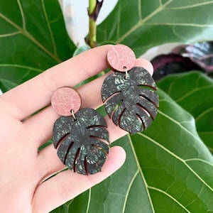 Woman holds green and pink monstera leaf earrings in-front of green house plant