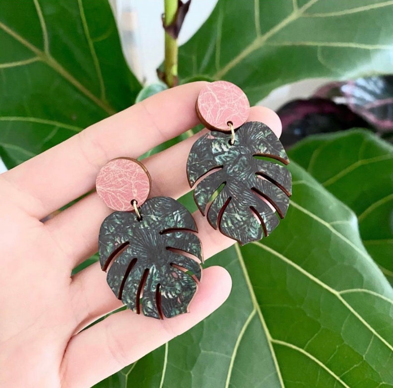 Woman holds green and pink monstera leaf earrings in-front of green house plant