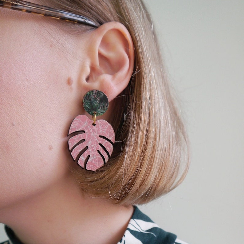 Pink and green monstera statement drop earrings hang from blonde women's ear