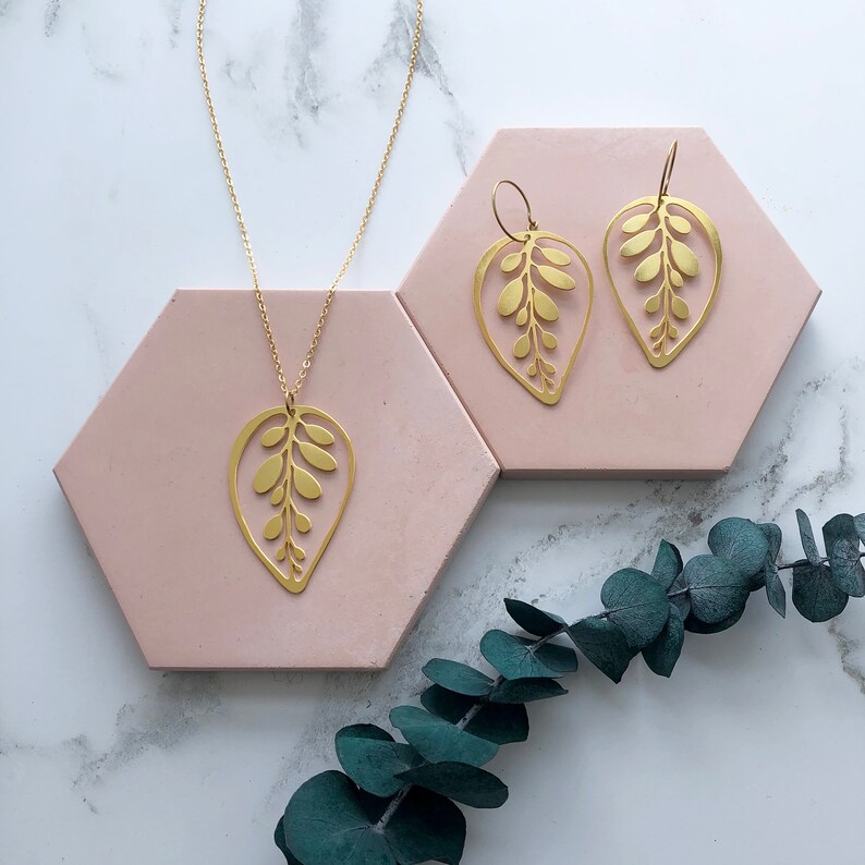 May include: A gold necklace and earrings set featuring a delicate leaf design. The necklace has a long chain and a pendant shaped like a leaf with intricate details. The earrings are also leaf-shaped and dangle from small hoops.