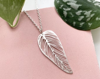 May include: A silver necklace with a delicate, leaf-shaped pendant. The pendant is cut out with a detailed design, creating a lacy effect. The necklace is displayed on a pink surface.