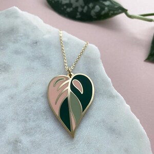 May include: A gold necklace with a pink, green, and dark green heart-shaped pendant featuring a leaf design.