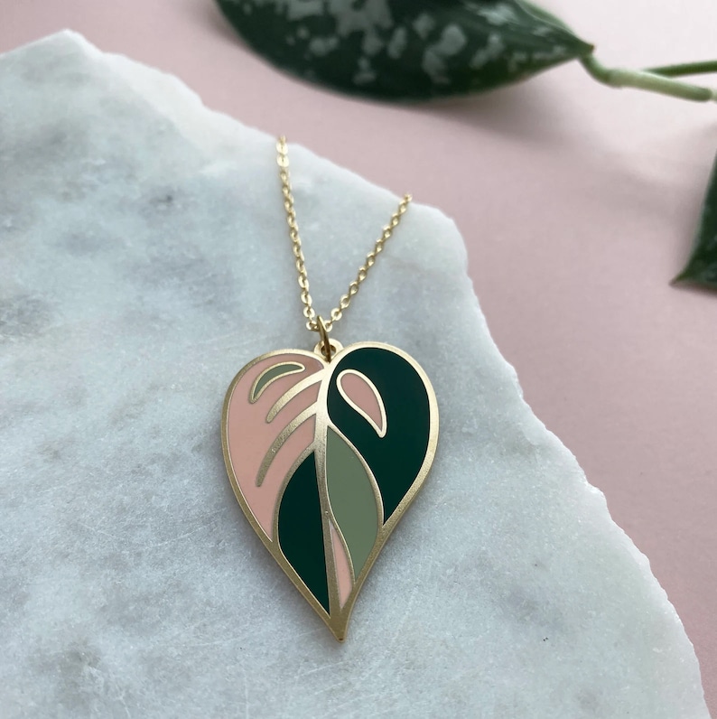 May include: A gold necklace with a pink, green, and dark green heart-shaped pendant featuring a leaf design.