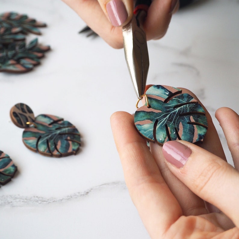 May include: A pair of earrings featuring a tropical leaf design with green and blue colors. The earrings are being assembled with pliers.