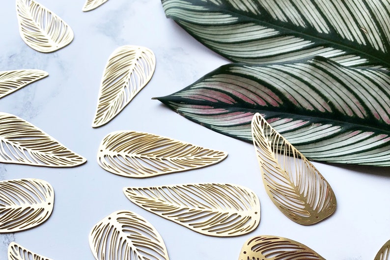 May include: A collection of gold leaf-shaped metal cutouts with intricate designs. The cutouts are arranged on a white surface with a green and white leaf in the background.