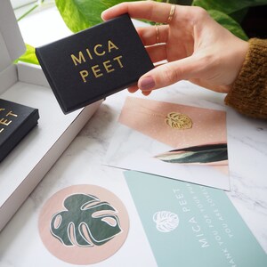May include: A black box with the text "MICA PEET" in gold lettering. The box is being held by a hand, and there is a white card with a gold leaf design and the text "MICA PEET" in the background.