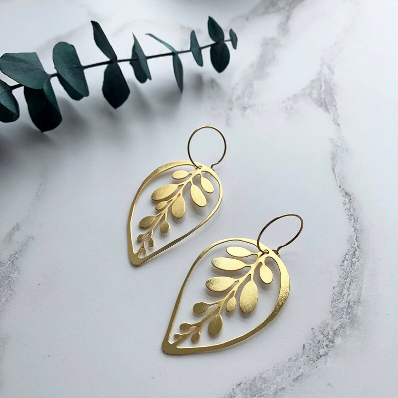 May include: A pair of gold leaf-shaped earrings with a delicate cutout design. The earrings are made of brass and have a simple, elegant style.