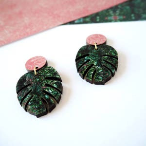 May include: A pair of green and pink monstera leaf earrings. The leaves are made of wood and have a green and pink floral pattern. The earrings are attached to a pink circle.