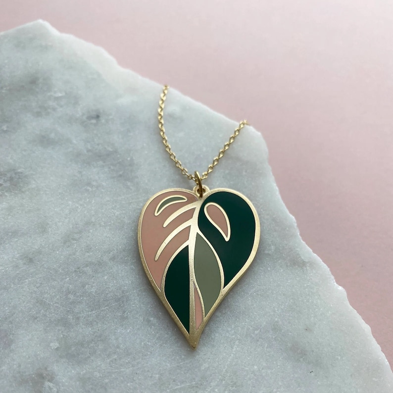 May include: A gold necklace with a heart-shaped pendant featuring a pink and green leaf design.