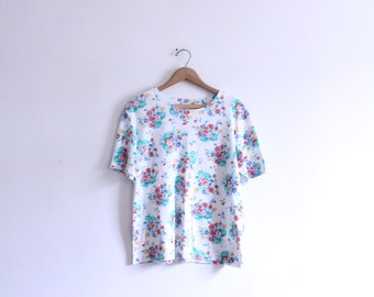 Puede incluir: Una camiseta blanca con estampado floral con flores rojas, azules y amarillas.