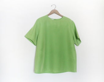 Puede incluir: Una blusa de manga corta de color verde claro colgada de una percha de madera sobre un fondo blanco.