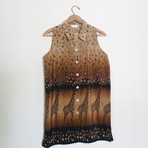 Op de afbeelding: Een mouwloos knoopshirt met een bruine en zwarte giraffenprint. Het shirt heeft een kraag en een knoopsluiting aan de voorkant.