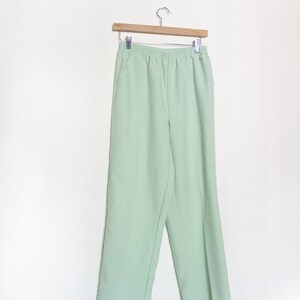 Pode incluir: Uma calça verde claro com cós elástico. A calça está pendurada em um cabide de madeira.