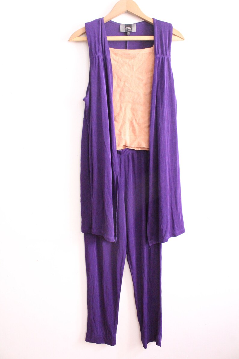 Puede incluir: Un conjunto de tres piezas en morado y melocot&oacute;n. El conjunto incluye una chaqueta larga sin mangas, un top sin mangas y pantalones. La chaqueta y los pantalones son de un morado intenso, mientras que el top es de color melocot&oacute;n.