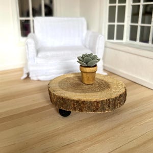 May include: Miniature wooden side table with a natural wood slice top, featuring a small potted succulent. The table has a rustic, natural aesthetic and is set in a dollhouse scene with a white armchair and window.