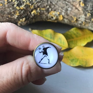 Puede incluir: Un anillo redondo de vidrio blanco cabujón con una imagen en blanco y negro de una persona lanzando una pelota. El anillo está engastado en un metal de color bronce.