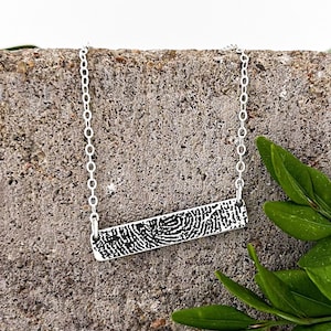 May include: A silver chain necklace with a bar pendant featuring a fingerprint design. The necklace is displayed on a textured gray surface with green foliage.