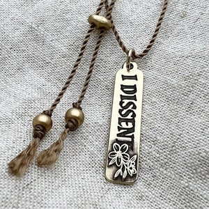 Brass pendant engraved with 'I DISSENT' on a textured fabric background
