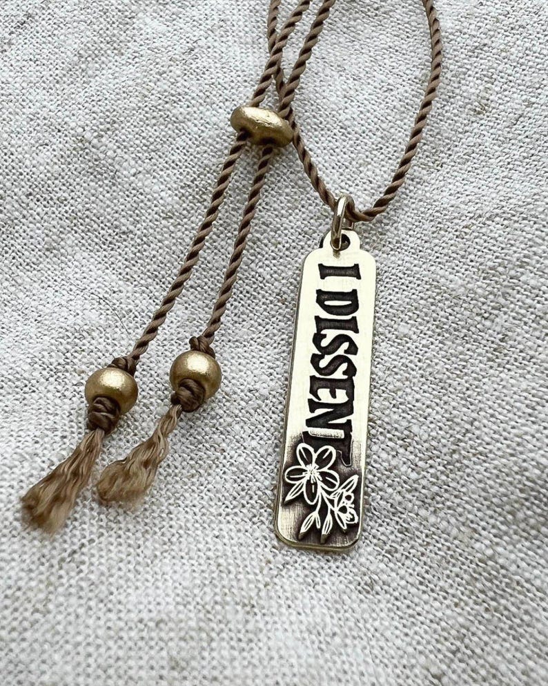 Brass pendant engraved with 'I DISSENT' on a textured fabric background