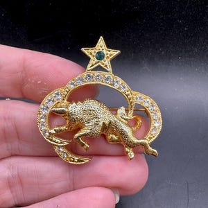 May include: Gold-toned brooch featuring a textured bull, a star with a green gem, and a ring adorned with clear crystals. The brooch has a pin on the back for secure attachment. A vintage-style accessory.